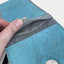 Hand, die die Innentasche einer handgefertigten Handytasche aus Filz und Baumwolle in Hellgrau und Azurblau präsentiert.