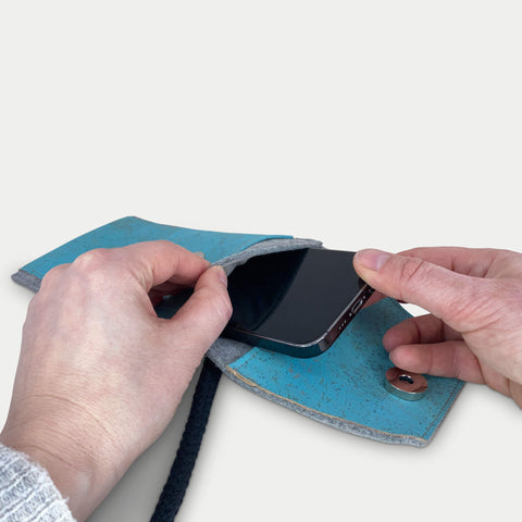 Person, die ein iPhone 16 Pro Max in eine stilvolle hellgraue und blaue Filztasche mit Schulterriemen steckt.